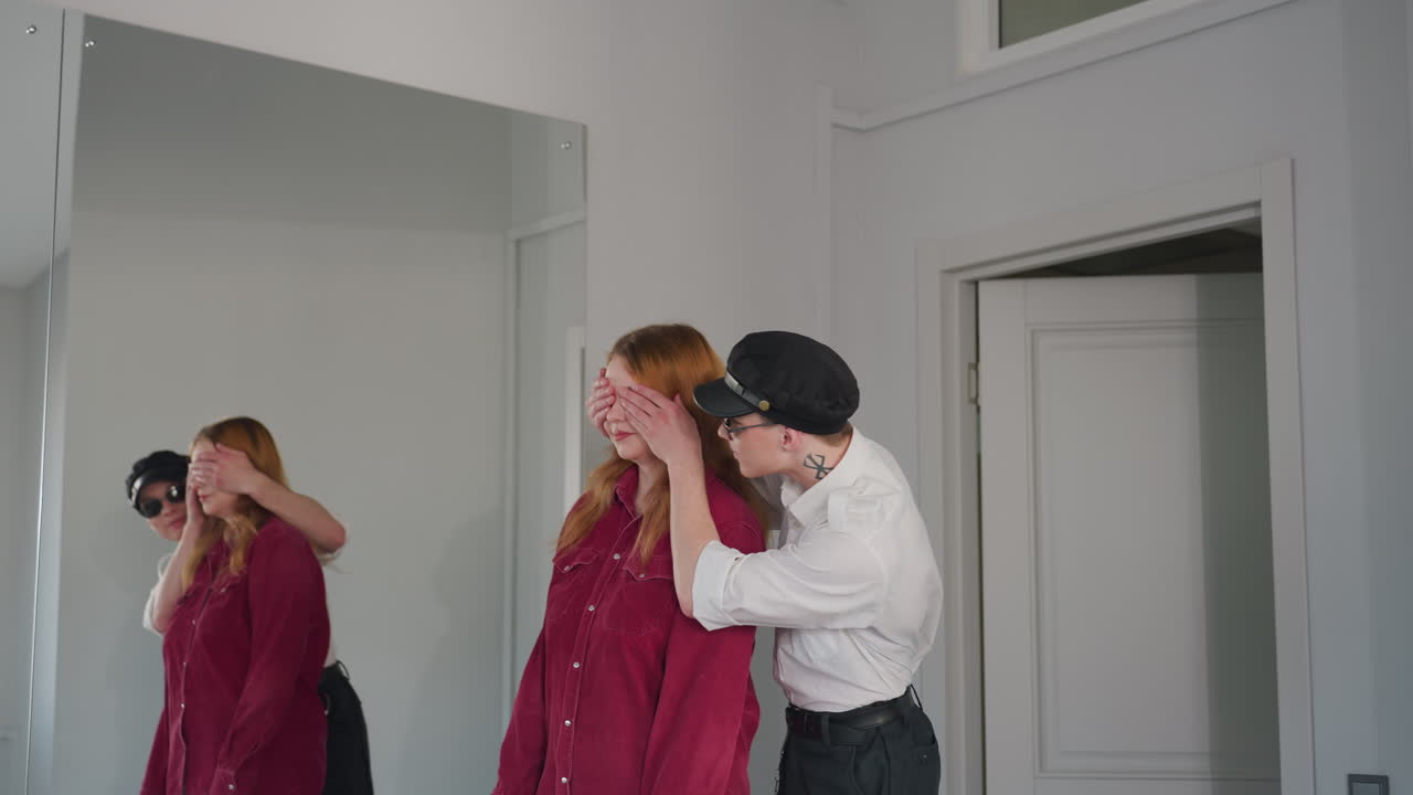 Young man in cap and uniform playfully covers eyes of unhappy woman in red shirt while gently leading her toward seat, mirror on wall reflects tender yet awkward moment in bright interior space