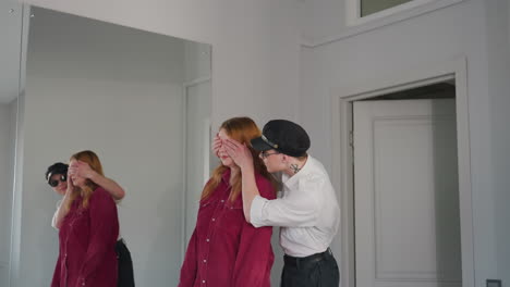 young man in cap and uniform playfully covers eyes of unhappy woman in red shirt while gently leading her toward seat, mirror on wall reflects tender yet awkward moment in bright interior space