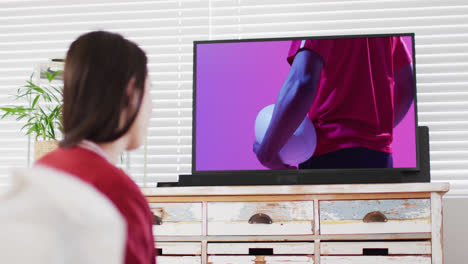 Caucasian-woman-watching-tv-with-african-american-male-rugby-player-with-ball-on-screen