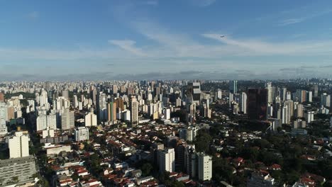 smooth-sideways-motion-with-drone-filming-a-sea-of-buildings-in-city-with-blue-sky,-sunny-day-with-plane-passing-by-in-the-background-in-4K-resolution