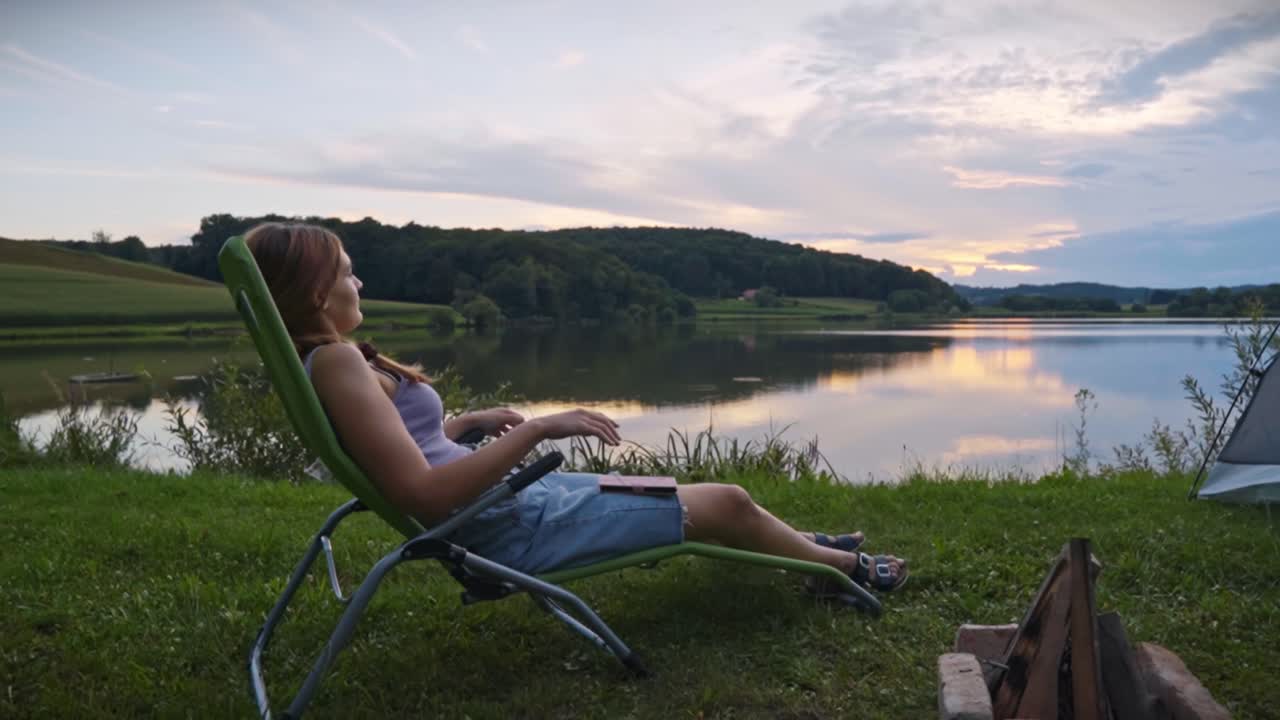 Frau liegt auf einem Liegestuhl nahe einem ruhigen See mit Lagerfeuer bei Sonnenuntergang, genießt die friedliche Campingatmosphäre und die malerische Naturlandschaft in Savci, Slowenien.