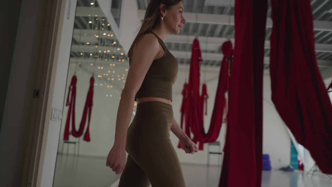 Well lit yoga studio with transparent glass windows and lights illuminating interior as aerial yogi walks through door clad in fitted activewear with red hammock straps hanging in background