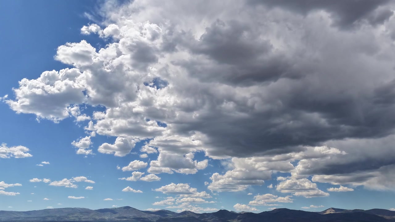 4K drone timelapse captures dark, brewing storm clouds developing over a rugged mountain range. Cinematic aerial motion reveals high-contrast weather with clear blue skies in the same wide vista.