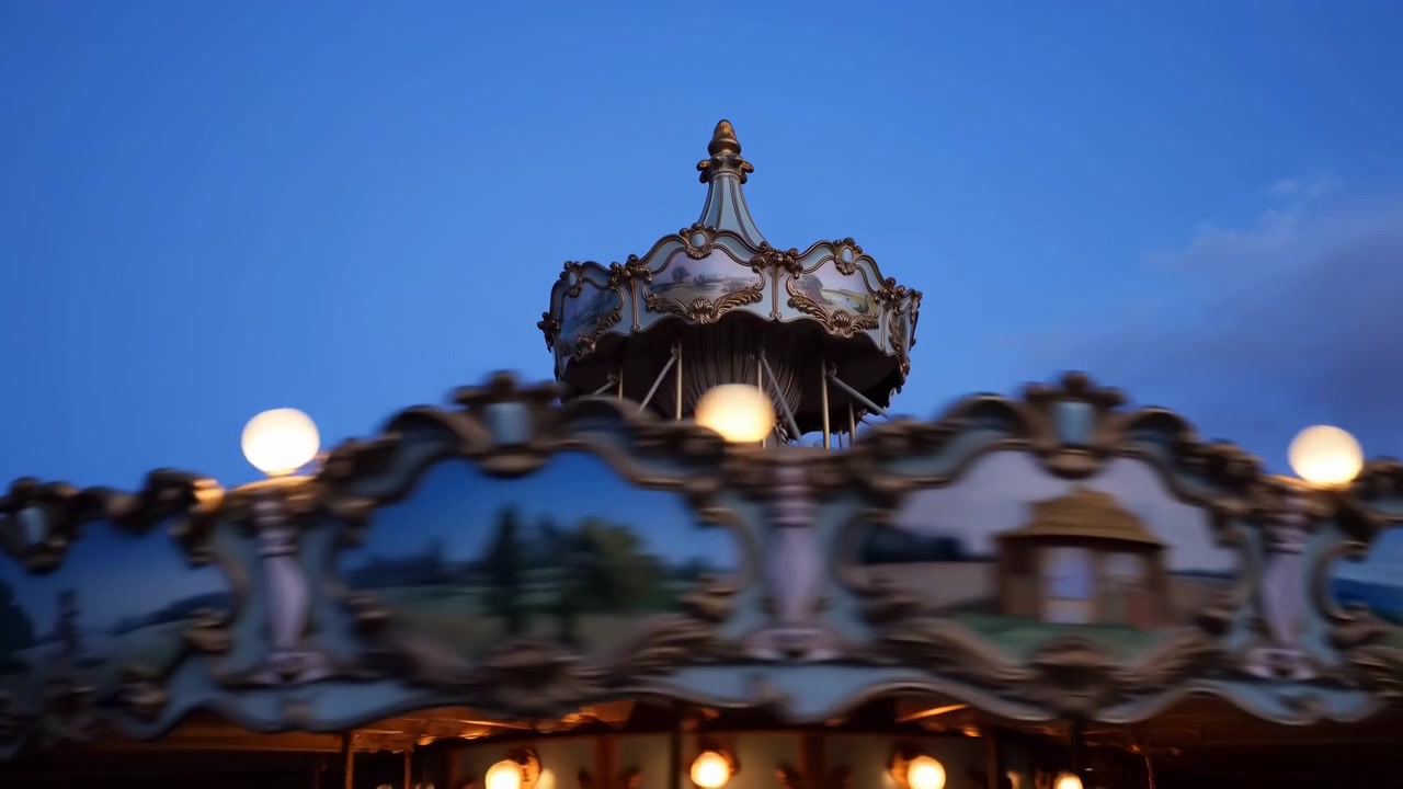 Close-up view of a rotating vintage carousel canopy captured at dusk, focusing on an ornate painted crown turret and scalloped decorative panels. Warm round