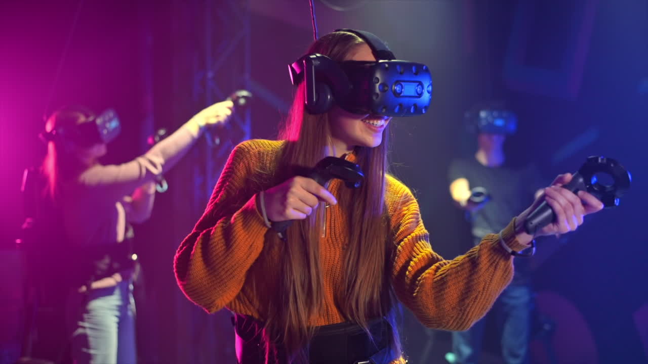 Una bambina felice e interessata gioca su un tapis roulant VR con casco e controller. Altre persone giocano nelle vicinanze. Slow motion