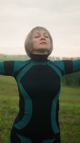 woman in green black suit stands with head slightly lifted, eyes closed, and hands slowly outstretched and brought down in a serene motion, practicing yoga in a vast open field under a cloudy sky
