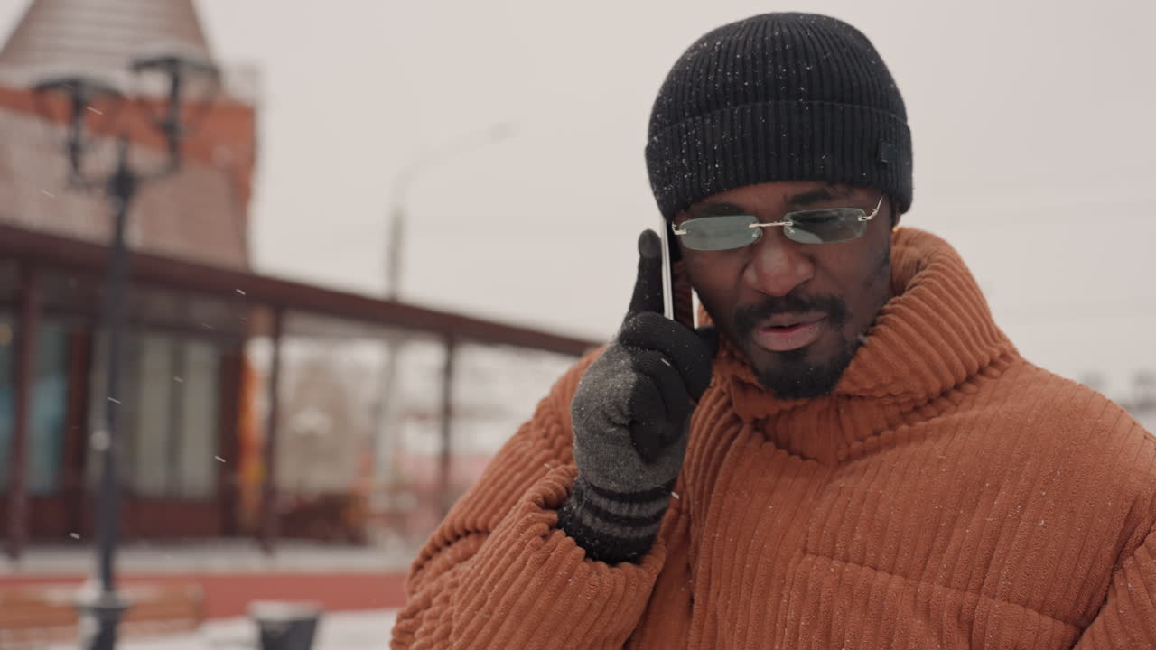 Black man talking on phone in brown knit coat and black beanie, wearing glasses and wool gloves, backpack over shoulder, walking through snowy city street with falling snow, expressive smile, candid