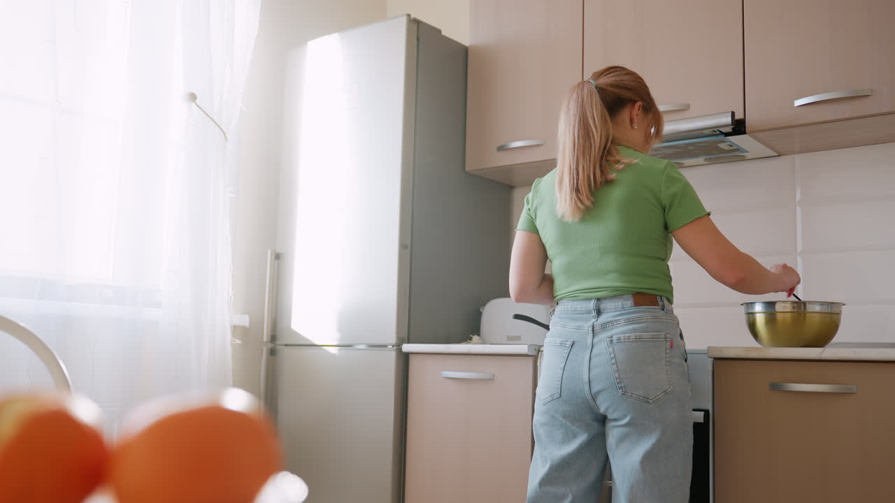 Back view of woman in green shirt and jeans stirring flour mixture in metal bowl on kitchen counter while oranges sit blurred in foreground with bright sunlight through window