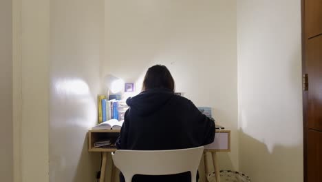 static wide shot of girl with black hair in jacket sitting and studying at desk