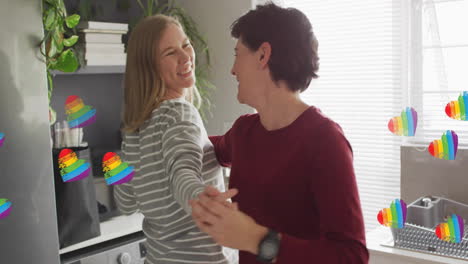 animation of pride rainbow hearts over happy caucasian lesbian couple dancing in kitchen