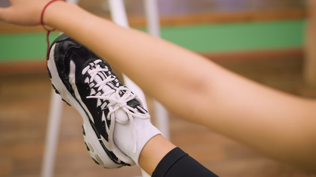 Girl places leg on bar stretching forward with extended hand while blurred face remains in background, black sneakers and white socks visible, exercise emphasizes balance, strength