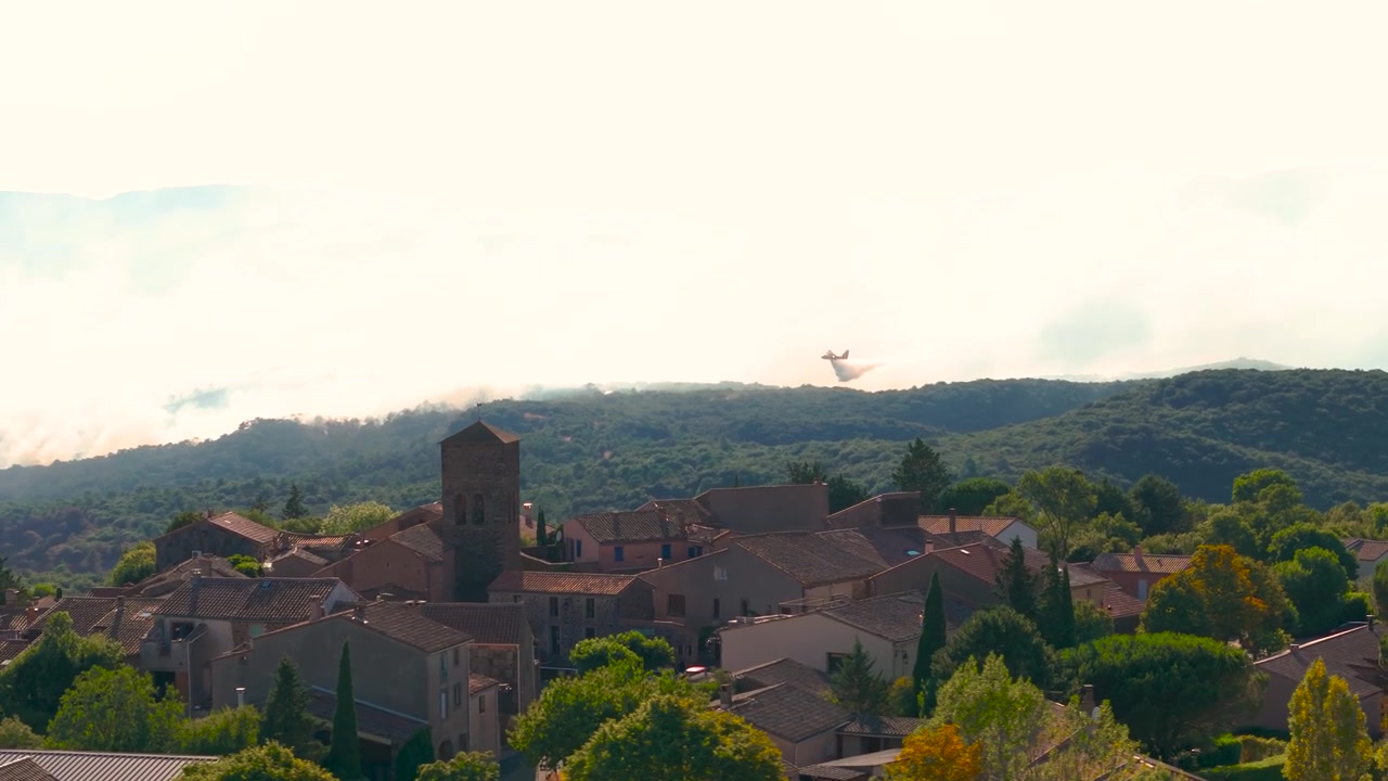 Riprese cinematografiche con drone a teleobiettivo che mostrano un aereo antincendio Canadair che vola attraverso un denso fumo in lontananza, giustapposto a un villaggio storico in pietra in primo piano, creando una prospettiva compressa