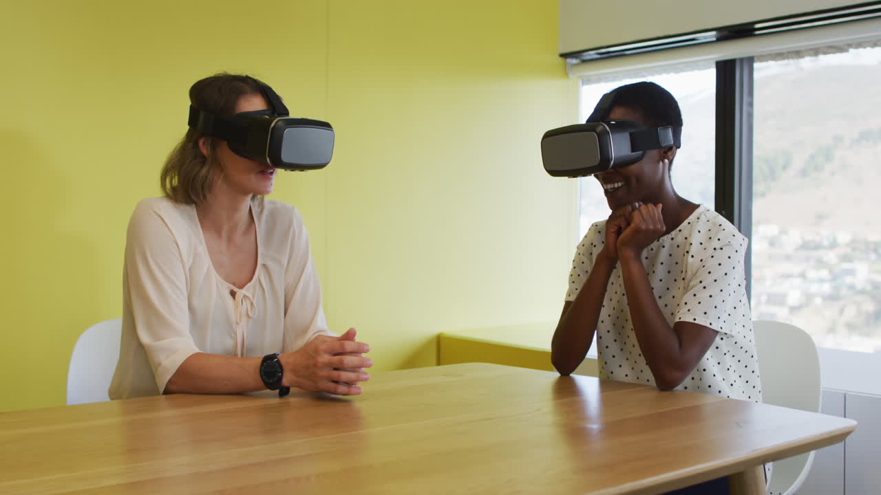 Two diverse female coworkers sitting at desk, testing vr googles