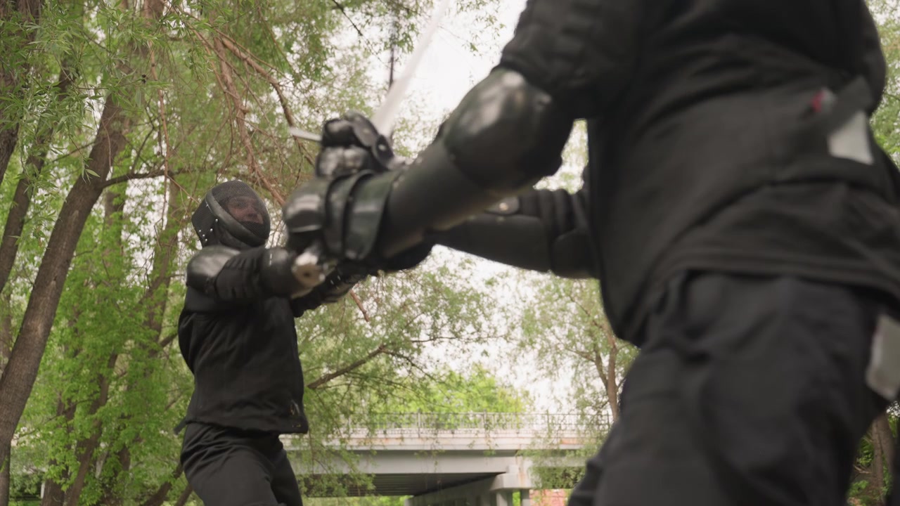 Two men in black protective gear engage in sword fight outdoors demonstrating various stances and movements with long bladed weapons. scene features dense green foliage and overcast sky