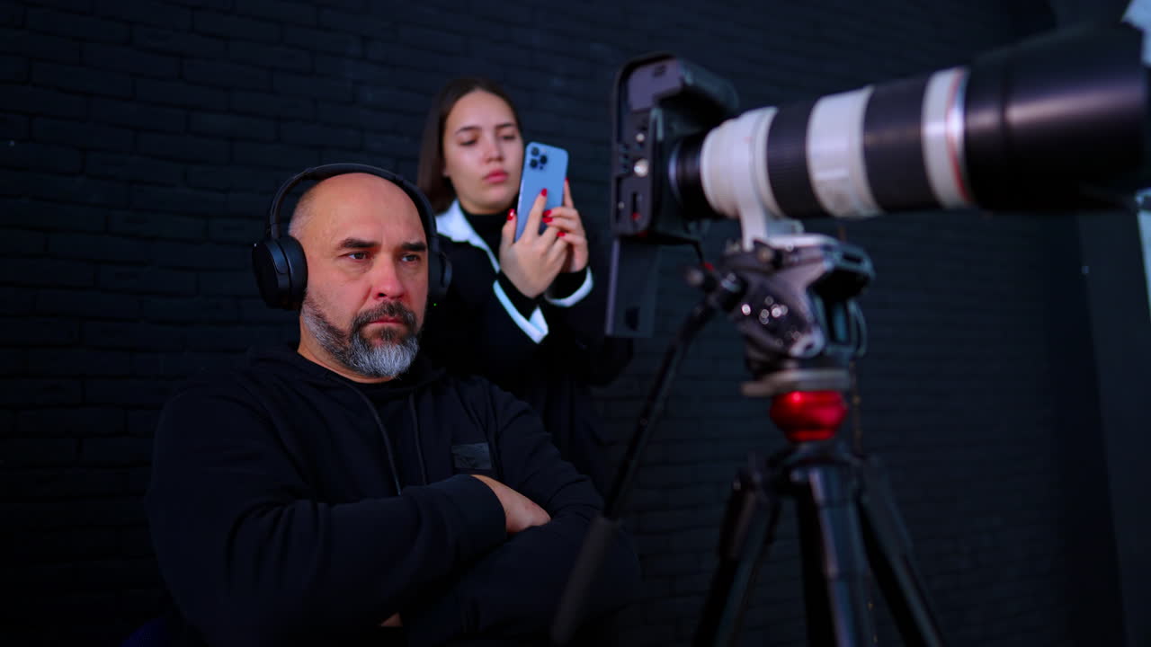 Ingegnere del suono con cuffie che monitora l'audio in studio. Primo piano di un uomo con le cuffie con una donna che tiene uno smartphone vicino a un obiettivo fotografico professionale