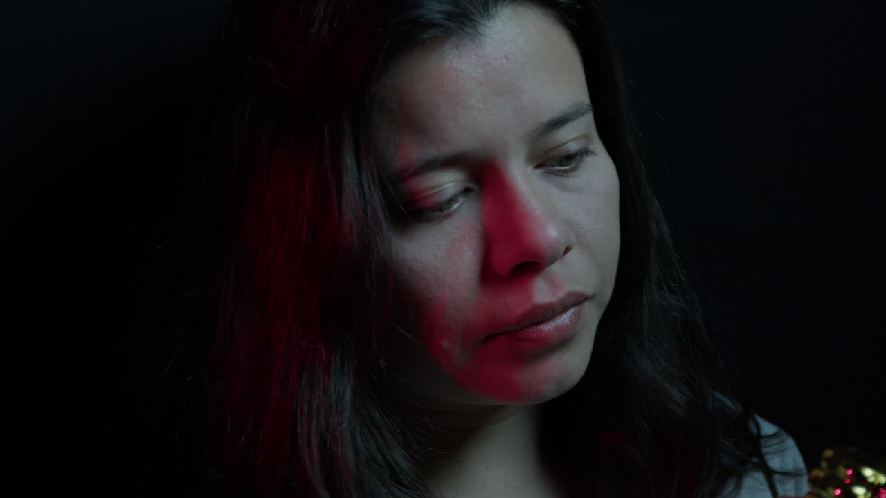 A young Hispanic girl holds a thumbs up award in a close-up shot. The scene features dramatic ambient lighting with red hues, showcasing her expression and the award's metallic texture