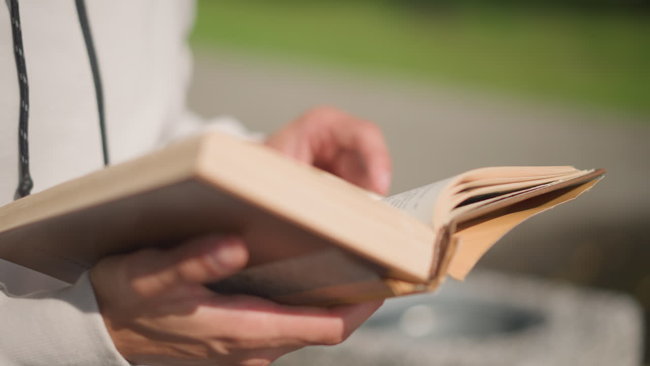 Primer plano de unas manos sujetando un libro abierto en un banco, con un parque iluminado por el sol de fondo; las páginas ondean suavemente con la brisa tenue y cubierta, papel agradable al tacto, lomo de libro de bolsillo antiguo, ambiente tranquilo y pausado ideal para estudiar