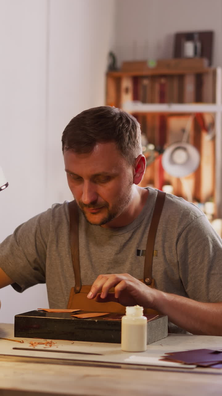 Leathersmith adjusts table lamp for comfortable leather processing in workshop. Bearded master crafts item in small studio. Workplace of handcraft designer