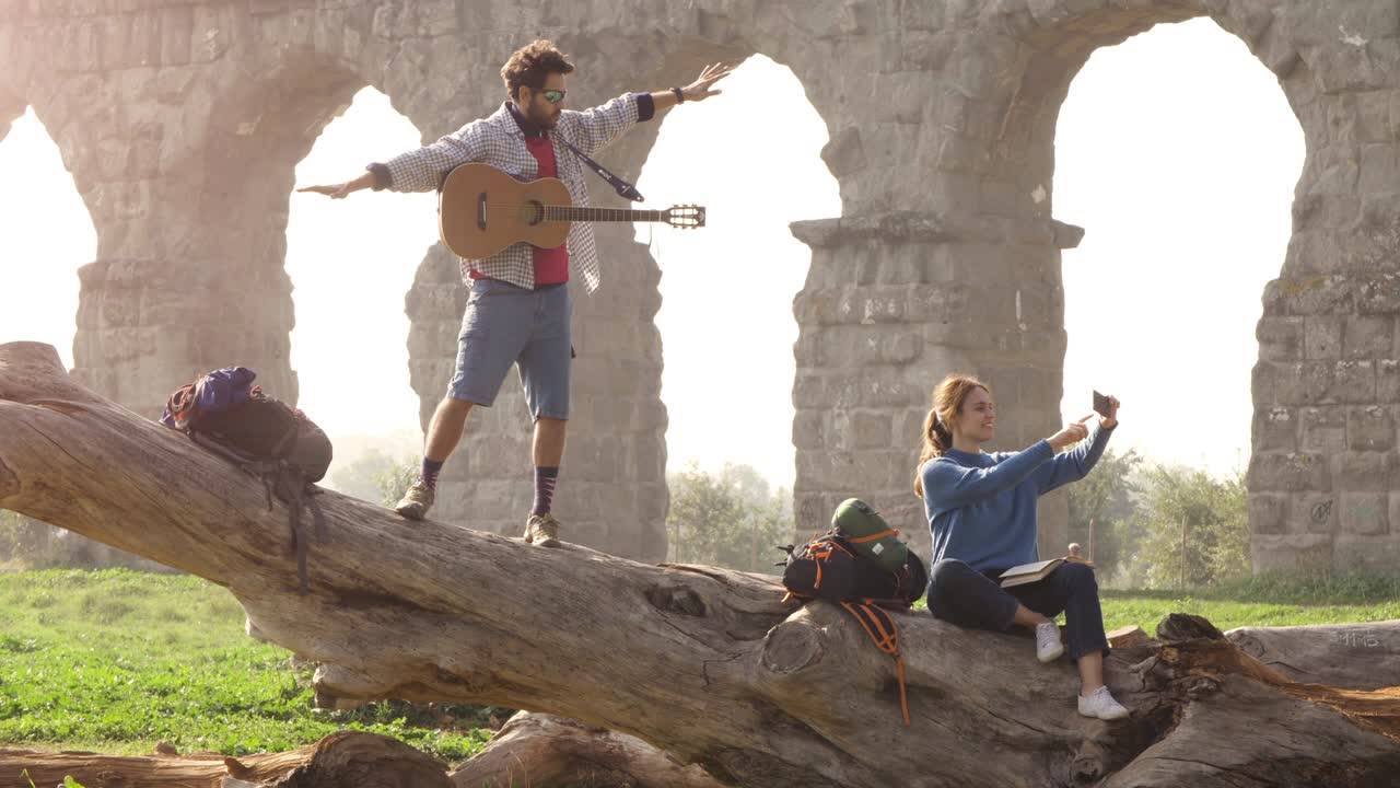 una pareja feliz de mochileros turistas en un tronco de troncos tomando selfies fotos con el teléfono inteligente frente a las antiguas ruinas del acueducto romano en el romántico parque parco degli aquedotti en roma al amanecer brumoso trípode de cámara lenta