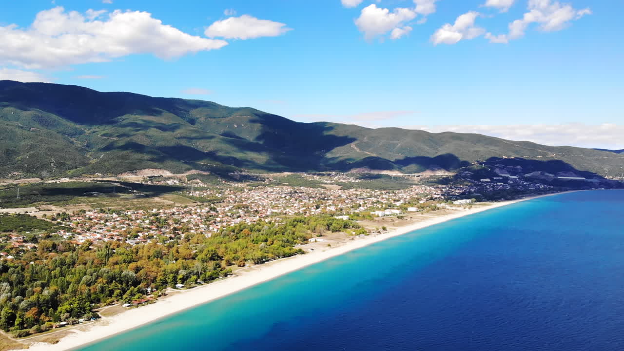 Panorama of the Asprovalta located on the Aegean sea coast with buildings and greenery, green hills. Long beach along the town. Sunny day, view from the drone. Greece