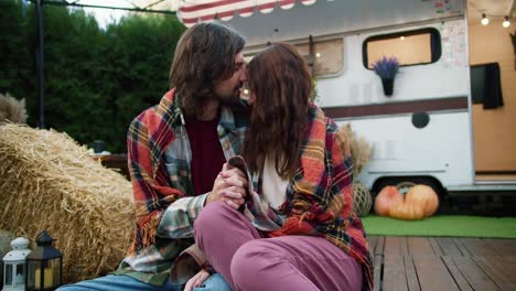 Close-up-shot-of-a-happy-couple,-brunette-guy-in-a-green-checkered-shirt-sits-wrapped-in-a-red-checkered-blanket-with-his-brunette-girlfriend-near-their-trailer-in-a-camp-outside-the-city-during-a-picnic-in-the-summer