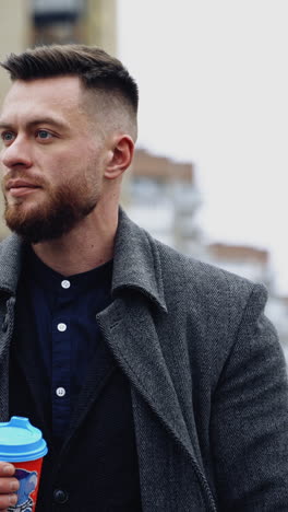 portrait of a smiling businessman outdoors. young bearded man holds a plastic glass of coffee in the city street and looks on camera merrily. vertical video