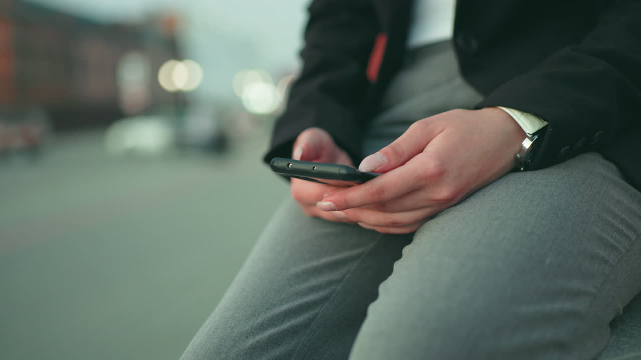 Close up of girl using smartphone while seated in formal wear, soft bokeh background with indistinct objects moving in urban setting, fingers interacting with phone screen in natural daylight