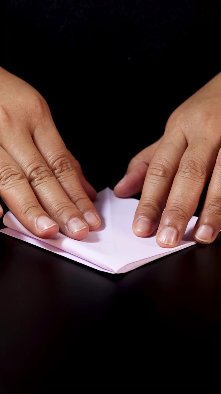 Adult hands carefully fold white paper on dark surface under soft, even lighting, close-up view