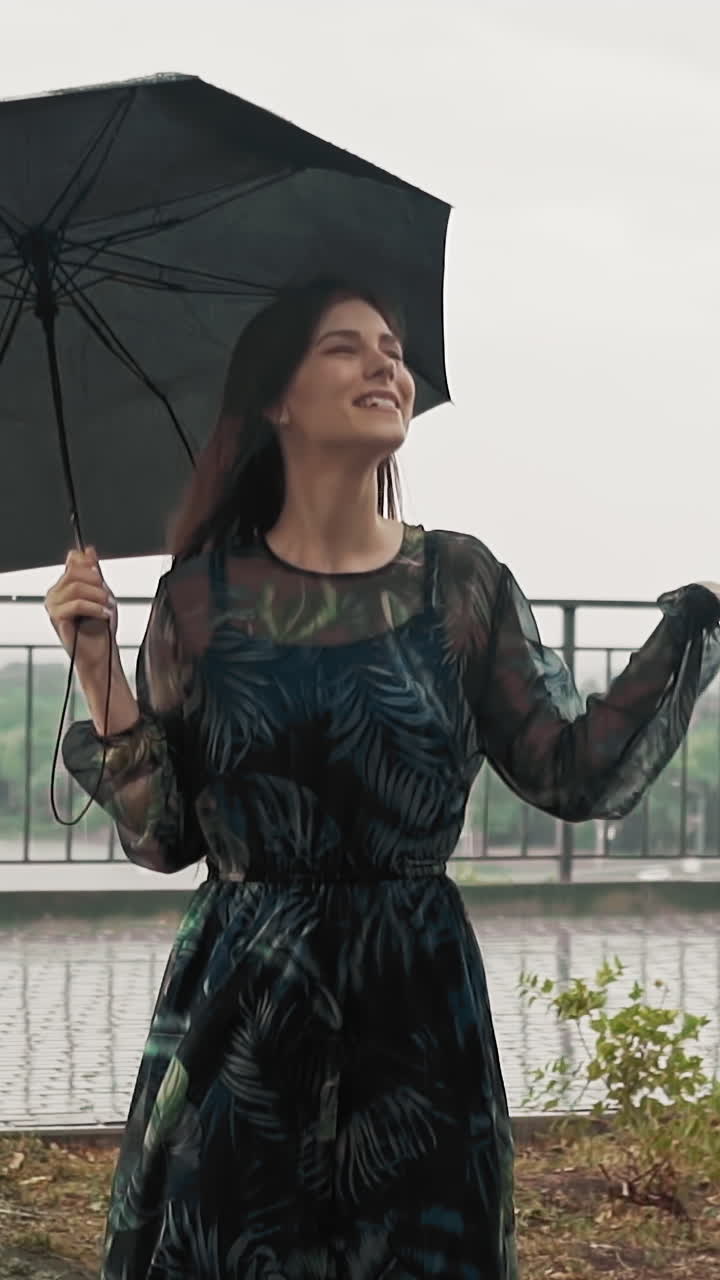 Positive young woman in elegant dress hides under black umbrella enjoying rainy weather on viewing deck on warm summer day slow motion