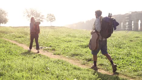 una joven pareja de mochileros turistas caminando cerca del acueducto romano arcos en el parco degli aquedotti ruinas del parque en roma al amanecer con guitarra y saco de dormir cámara estacionaria de cámara lenta