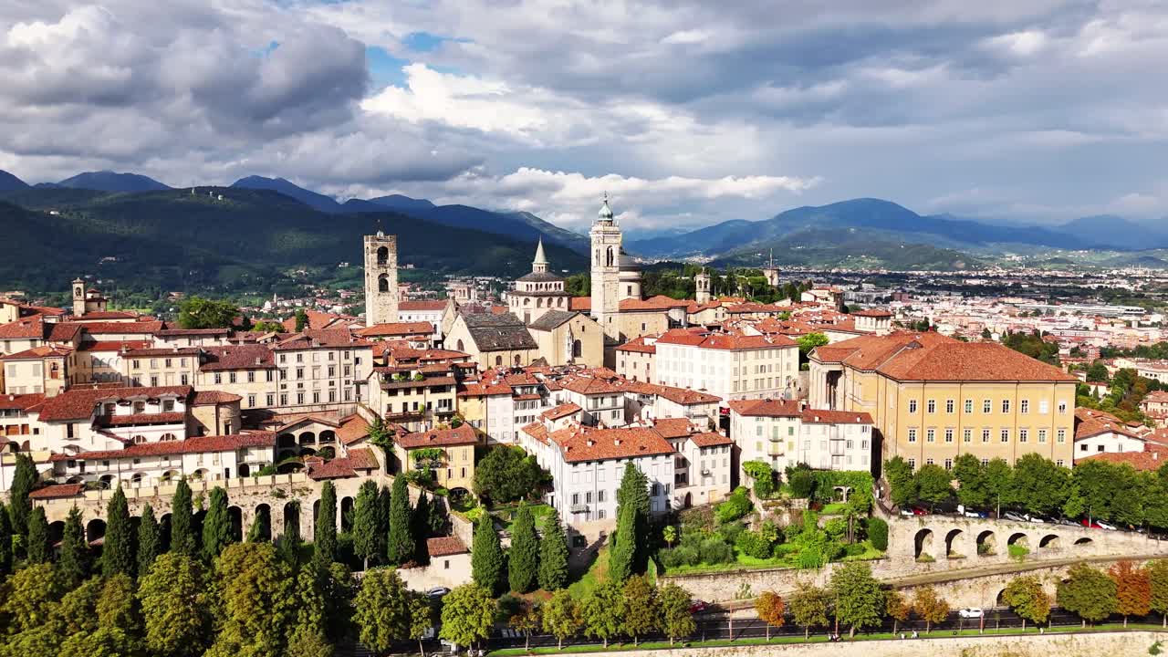 Una vista dettagliata di Bergamo, in Italia, con la sua architettura storica, i tetti in terracotta e le verdi colline incorniciate da montagne in lontananza sotto un cielo luminoso, che mette in risalto il paesaggio e l'atmosfera della città