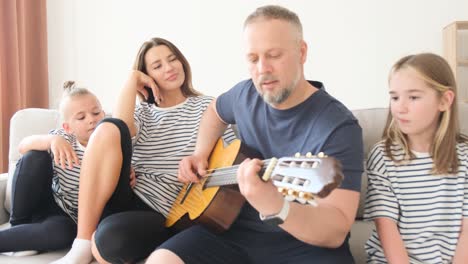 young dad playing guitar for his family while sitting on sofa. relaxing at home on the sofa, the husband sings for his wife. happy family relaxing together at home
