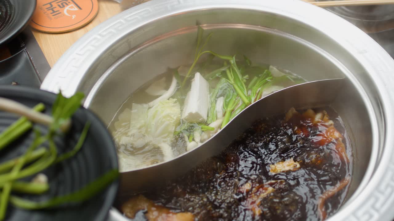 Fresh vegetables placed into steaming dual broth hotpot, overhead view, natural lighting, minimal camera movement