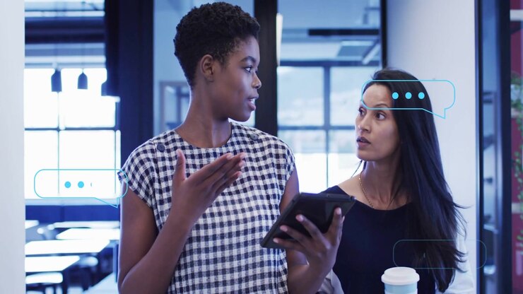 Left woman showing tablet with blue icons and tapping coworker leaning in reviewing business app