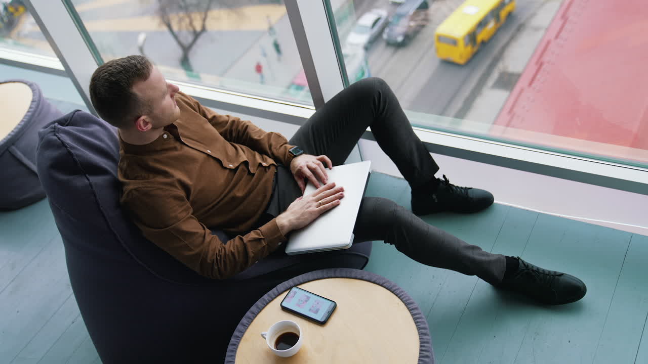 Tired businessman is having a break. Young man closes the laptop and looking through the window on a city landscape. Freelancer works in comfortable place.