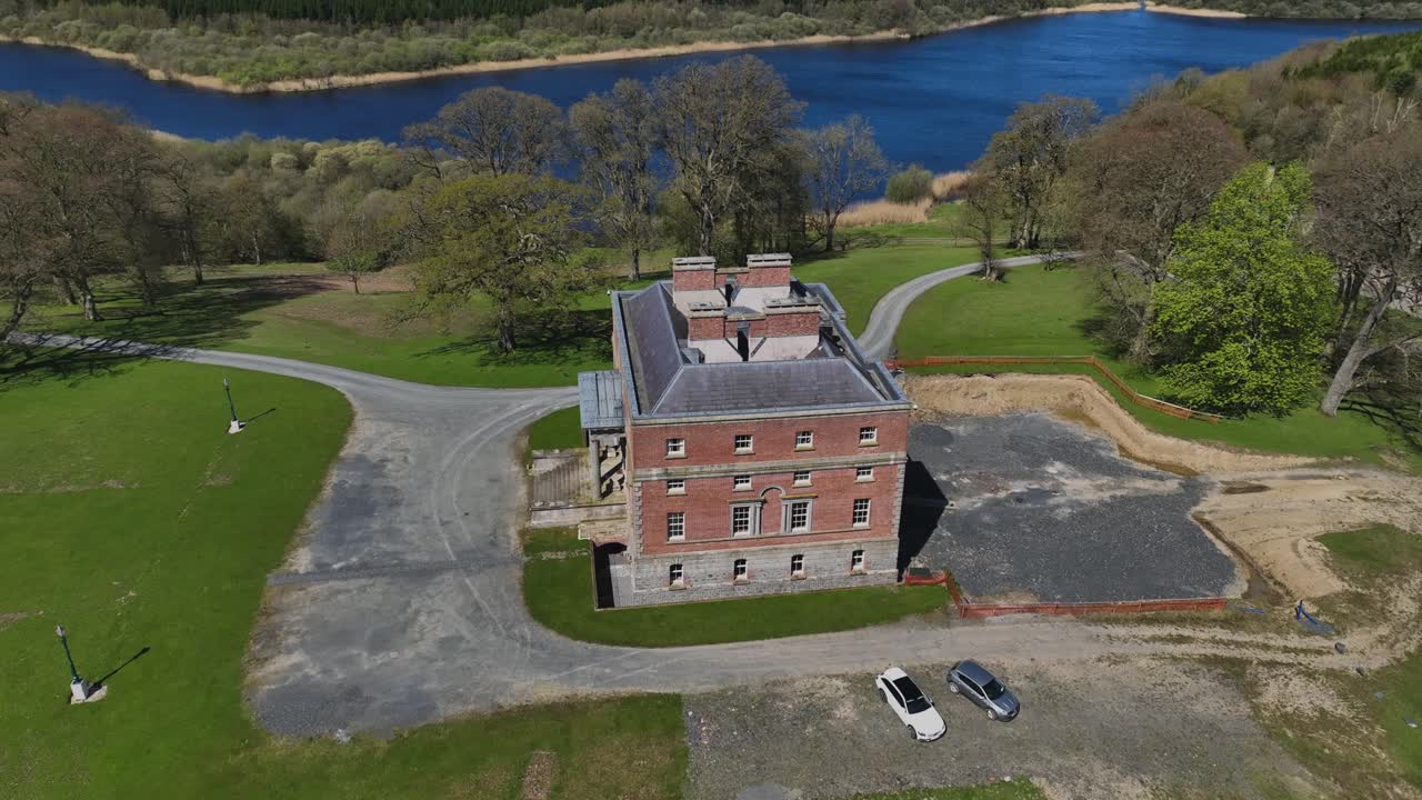 Bellamont House, County Cavan, Ireland, April 2023. Drone circles red brick designed historic Georgian mansion, estate lawn, lake, trees and clear skies on sunny day.