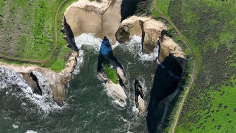 Top-down-view-of-coastline-shore-shark-fin-cove,-green-foliage-covering-rocks-against-waves-crashing-,-California