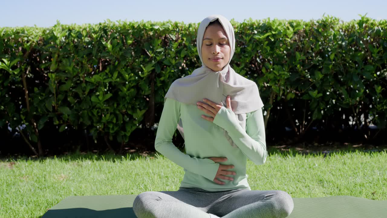 Wanita dewasa duduk bersila di atas matras hijau di halaman rumput yang cerah, mengenakan hijab, bernapas untuk menenangkan diri.