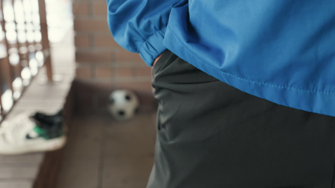 primer plano de chaqueta azul con balón de fútbol en el porche, jugador de pie con la mano en el bolsillo, tacos húmedos por la lluvia en el banco, pared de ladrillo de fondo, luz tenue, tensión y expectación prepartido