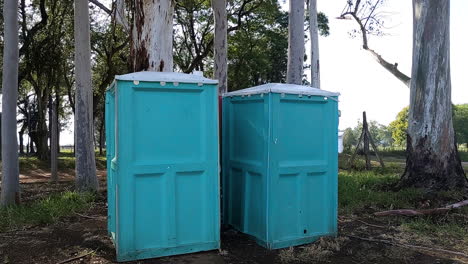 Carro-Delante-De-Dos-Baños-Portátiles-Azul-Verde-Agua-En-Un-Parque-Familiar-Con-árboles