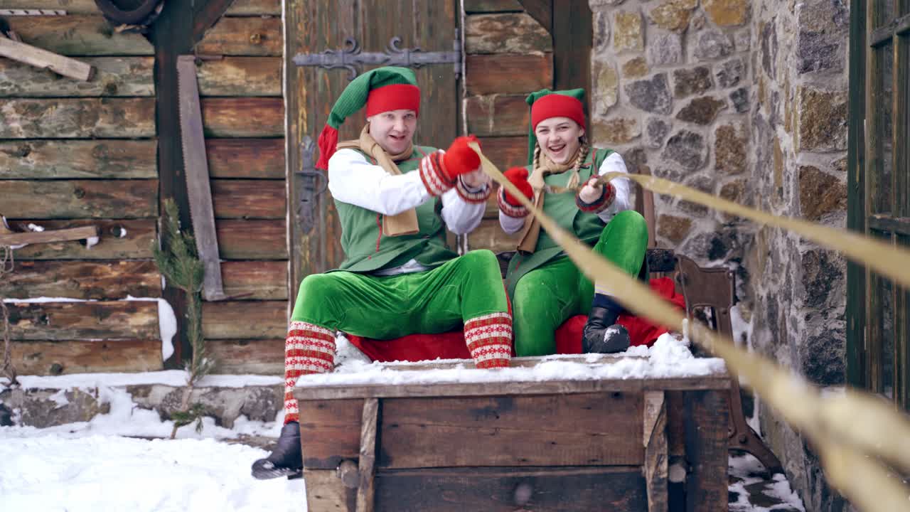 Two elves in green costumes sitting in wooden sleigh outdoors. Happy elves sit together in old fairy sleigh on the house background in winter. Christmas time.