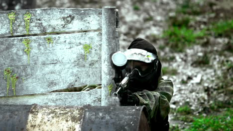 hombre disparando su arma de paintball agachado detrás de la valla