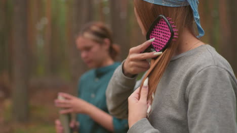 close-up of woman brushing her hair, focusing on her long reddish-brown hair and gentle self-care moment, while another woman in the background, slightly blurred, opens her thermos