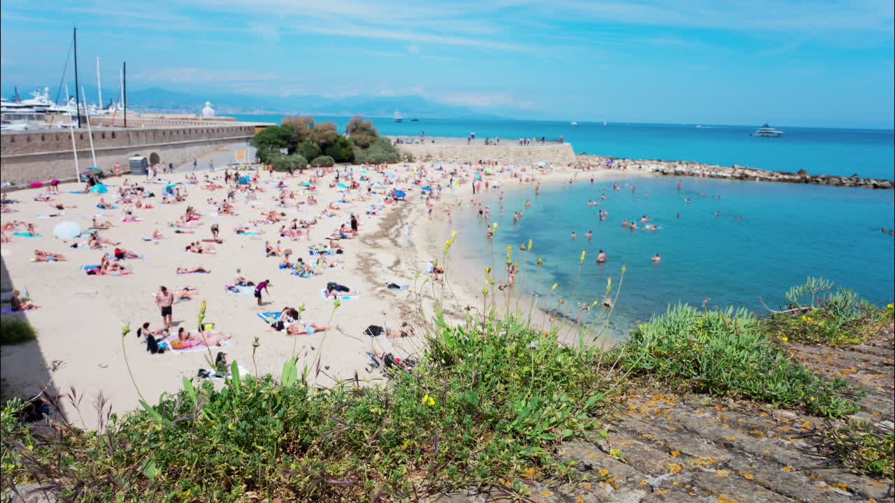 Antibes, France - May 17, 2025: High angle view of people relaxing on the Gravette beach with The Nomad urban sculpture by Jaume Plensa on the coast of the city in the background
