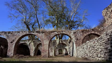 sunlight illuminates the ruins of the hacienda de san jacinto ixtoluca in morelos, mexico, with bare trees growing through the arches and a clear blue sky above, drone shot