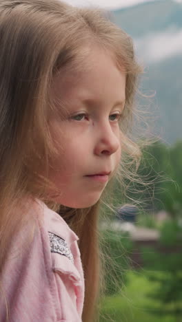 pretty little girl with long loose blonde hair stands near pine tree in hotel yard against large cloudy mountains close side view slow motion