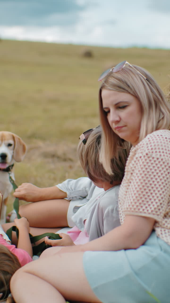머리에 선글라스를 입은 사려 깊은 여자가 근처의 친구와 함께 웃고, 어린 소녀는 읽고, 개는 열린 시골의 풀 근처의 배경에 서 있습니다.