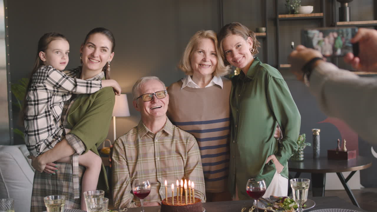 노인과 그의 가족은 집에서 생일 파티를 하는 동안 휴대폰 카메라로 포즈를 취하고 웃고 있습니다.