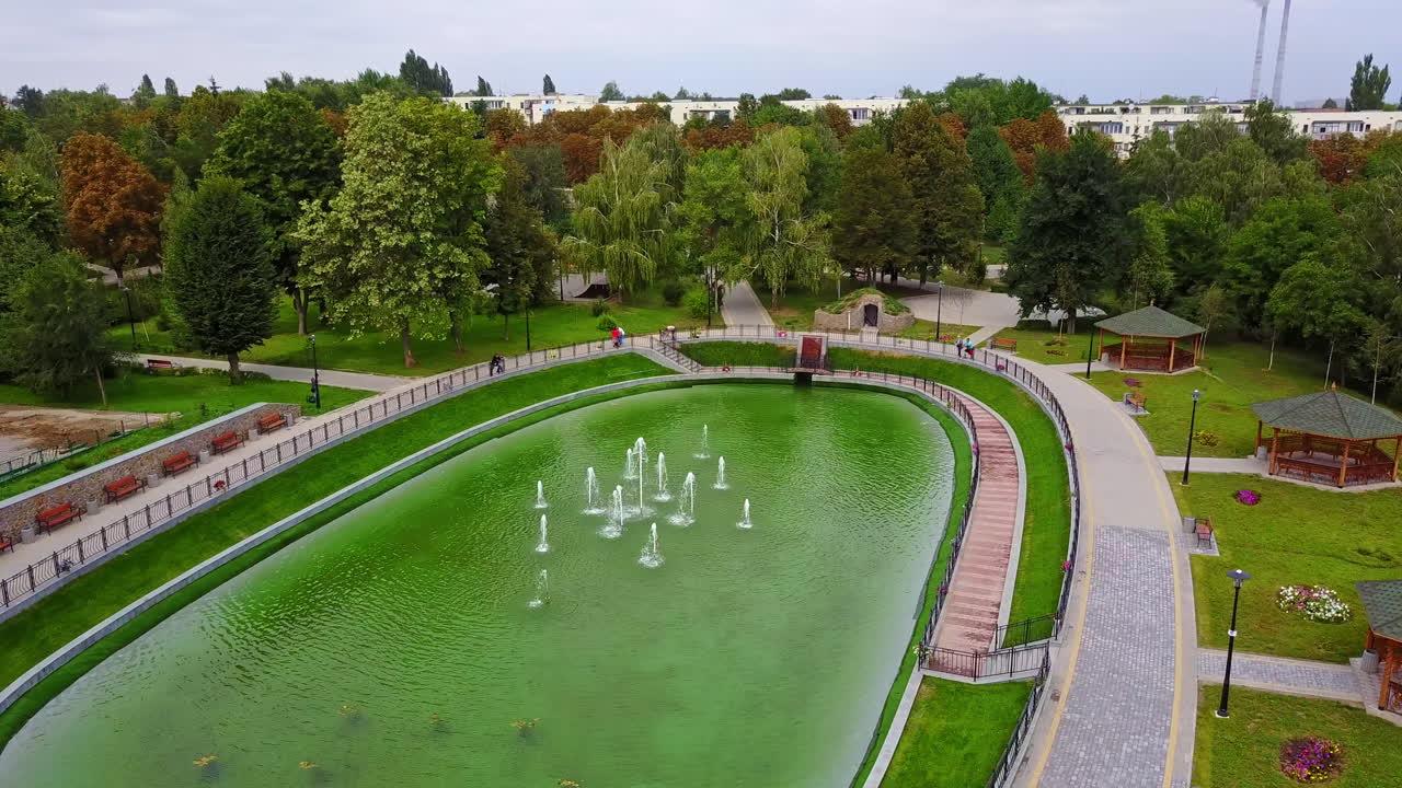 Oval pond with little fountains in the middle. Pavilions and benches for rest around the water. City park from top.