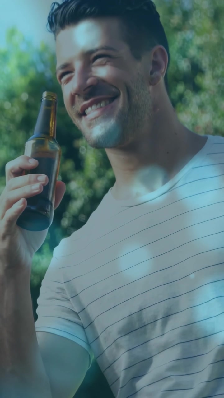 Cámara alejándose, hombre con camiseta a rayas levantando una botella marrón, bebiendo para refrescarse, bokeh en el jardín